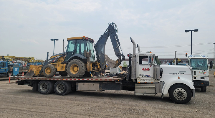 Heavy commercial flatbed truck performing equipment hauling by transporting a John Deere backhoe loader on a gravel lot.