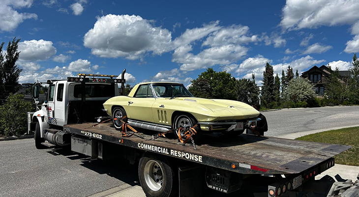 Commercial flatbed towing truck loaded with a vintage sports car parked on a paved residential road under a blue sky.